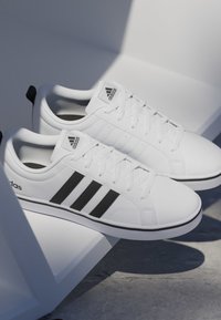 Par de zapatillas Adidas blancas con rayas negras y logo, exhibidas sobre una plataforma geométrica blanca contra un fondo gris.
