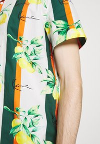 La camisa de manga corta presenta un tejido blanco con hojas verdes vibrantes y limones amarillos, acentuada por atrevidas franjas verticales en naranja y verde oscuro.