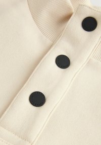 Top beige avec un col texturé à côtes, featuring trois boutons noirs avec un logo en relief, et des coutures plates pour une finition propre.