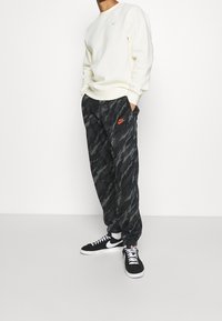 Svart- och gråmönstrade joggers med elastisk midja, orange Nike-logga, kombinerade med en cremefärgad sweatshirt och svarta sneakers med vita detaljer.