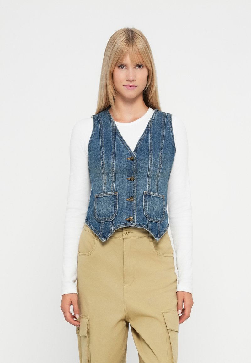 Τζην γιλέκο με V λαιμόκοψη, που διαθέτει χρυσά κουμπιά, δυο μπροστινές τσέπες και κατακόρυφη ραφή σε μπλε ύφασμα. Φοριέται πάνω από λευκό μακρύ μανίκι μπλουζάκι.