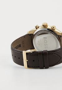 Reloj dorado con una correa de cuero marrón. Las características incluyen un fondo de caja transparente y botones de cronógrafo. Logo grabado en la correa.