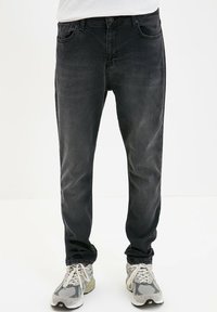 Jeans in denim nero con una vestibilità slim, dotati di cinque tasche, cuciture a vista e vita media. Indossati con scarpe da ginnastica grigie.