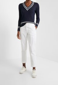 Granatowy sweter w splot kablowy z białym dekoltem w serek i paskami, zestawiony z białymi spodniami prostymi oraz kremowymi sneakersami z logo.