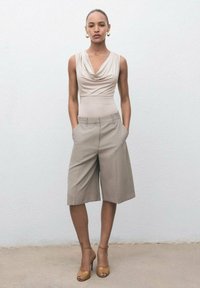 Top sin mangas de cuello de cowl beige, culottes neutros a medida y sandalias de tacón alto color tostado. La modelo posando con las manos en los bolsillos contra un fondo claro.