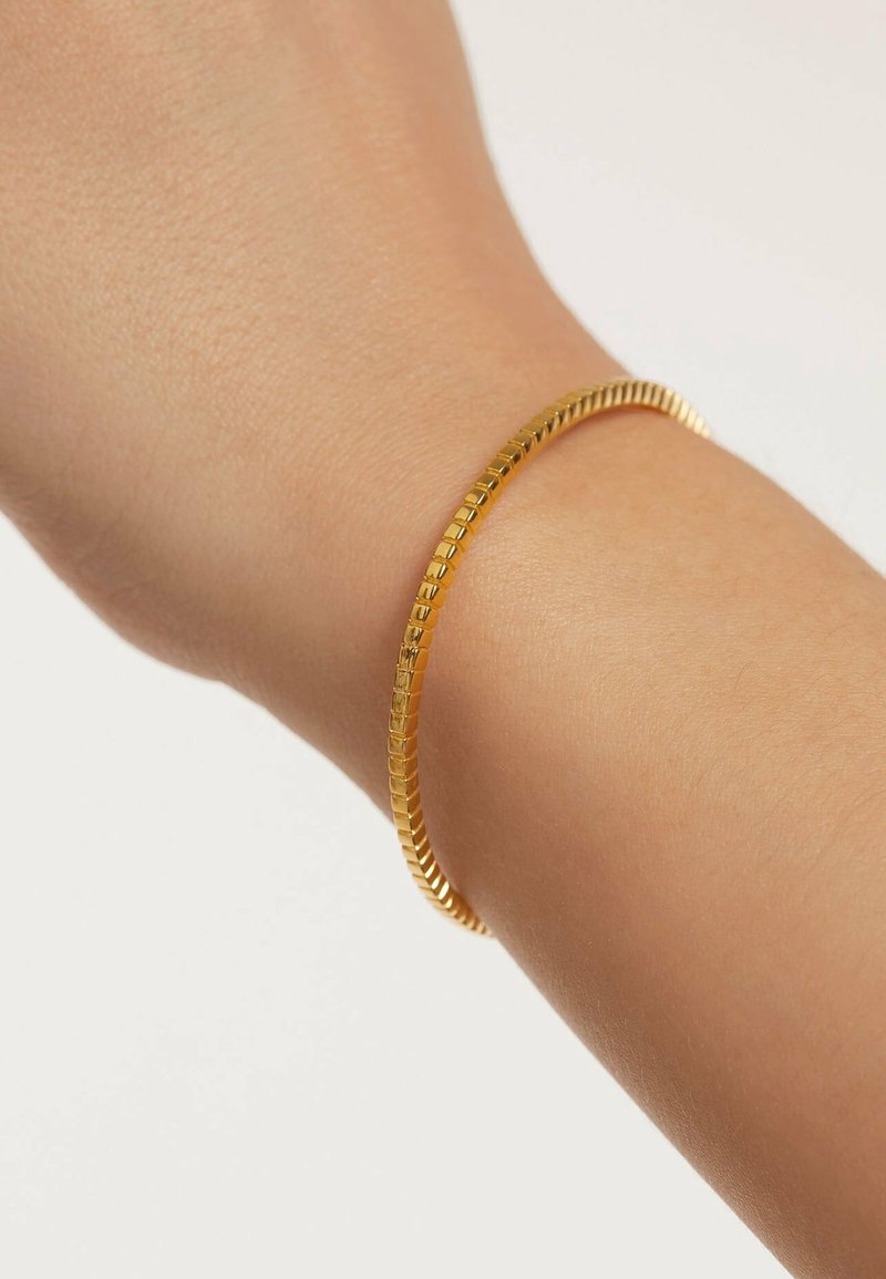 Bracelet fin en chaîne dorée porté au poignet avec une peau claire et lisse sur un fond clair uni.