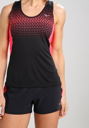Mujer con camiseta deportiva sin mangas negra y rosa y pantalones cortos negros, de pie delante de un fondo claro y liso.