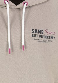 Beiger Hoodie mit weißen Kordeln, die mit pinkfarbenen Metallenden versehen sind, und gedrucktem Text: "SAME Same BUT DIFFERENT FRIENDSHIP IS ABOUT QUALITY NOT QUANTITY."