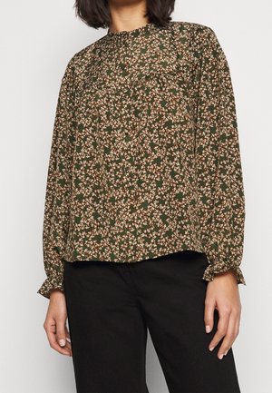 Femme portant un chemisier marron à manches longues avec un petit motif floral et un pantalon noir, debout devant un fond blanc uni.