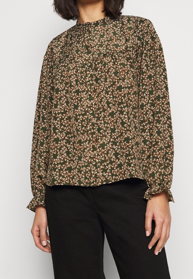 Femme portant un chemisier marron à manches longues avec un petit motif floral et un pantalon noir, debout devant un fond blanc uni.