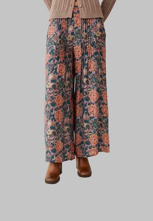 Persona que lleva pantalones de pierna ancha con estampado floral en rosa, verde y beige, botas marrones con tacón y una chaqueta de punto beige.