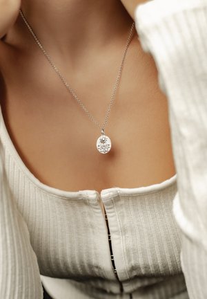 Silver pendant necklace on a fine chain featuring an oval shape with floral engravings, displayed against a textured white top.