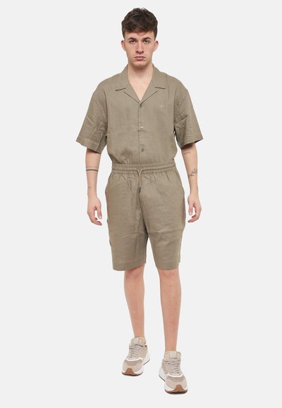 Camicia khaki a maniche corte con colletto e bottoni, abbinata a pantaloni corti coordinati. Tessuto leggero e testurizzato; cintura elastica; indossato con sneakers casual.