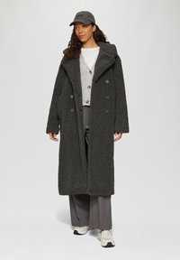 Donkergrijze, lange, fluffy jas met een brede kraag, dubbelzijdige knopen, gedragen over een lichtgrijze cardigan en grijze broek, gecombineerd met sneakers.