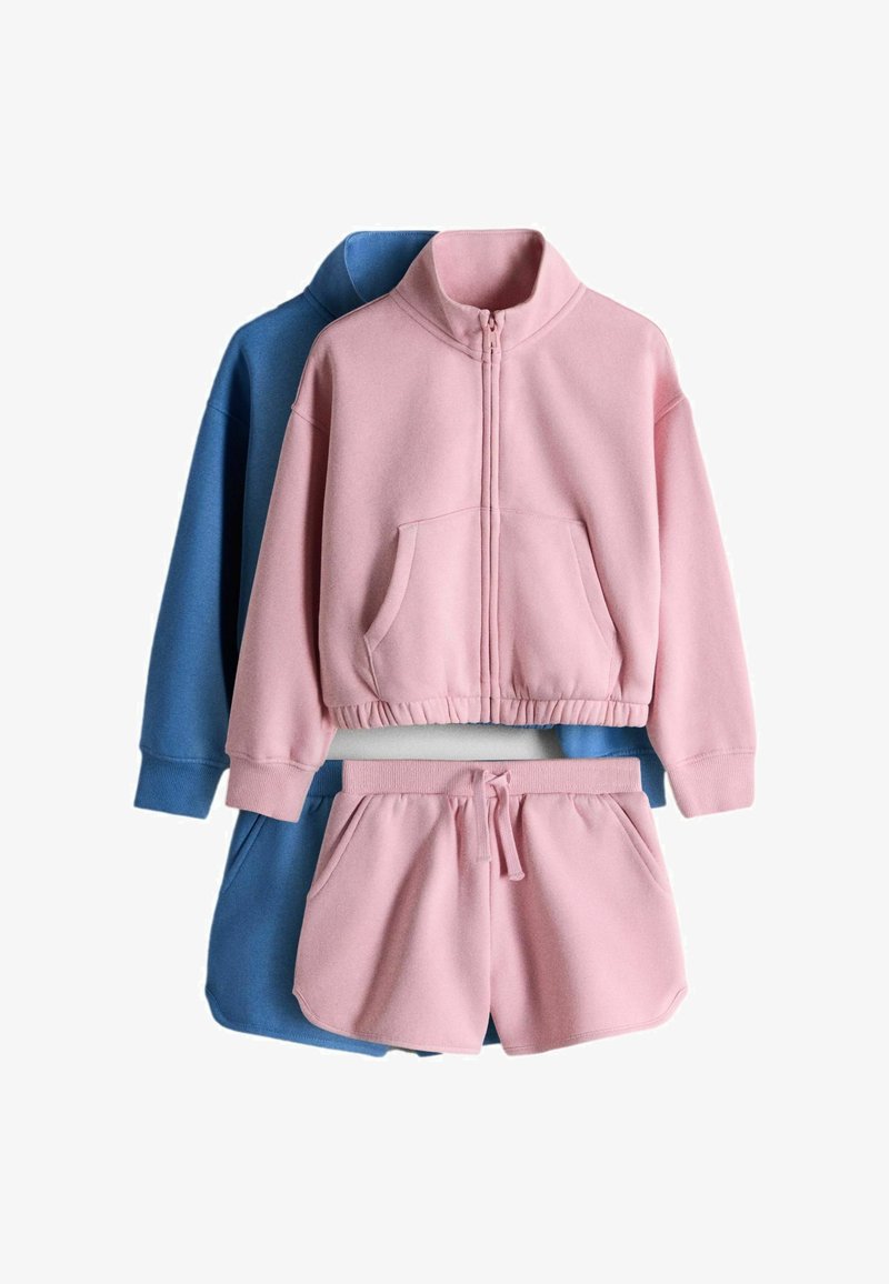 Roze fleece jas met rits en kangoeroezak, gecombineerd met bijpassende shorts, gedragen over een blauw fleece shirt met lange mouwen en shorts.