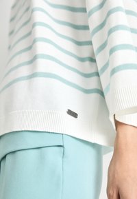Pulover alb cu dungi bleu deschis, manșete ribbate și un logo argintiu la tiv. Asortat cu pantaloni bleu deschis.
