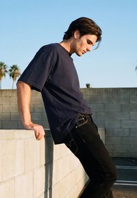 Noor mees mustades teksades ja tumedas t-särgis loivamas tagasi betoonseina vastu õues, taustal palmid ja selge taevas.