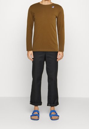 Camisa marrón de manga larga con un pequeño logo, combinada con pantalones negros y sandalias azules. Zapatos planos con doble tira y suela texturizada.