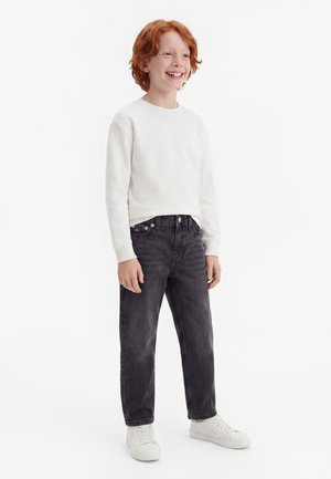 Enfant roux souriant debout, portant une chemise blanche à manches longues, un jean noir et des baskets blanches sur un fond blanc uni.