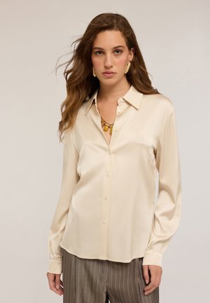Vrouw met lang golvend haar, gekleed in een beige zijden overhemd met knopen, gouden ketting, gouden oorbellen en gestreepte broek, tegen een lichte achtergrond.