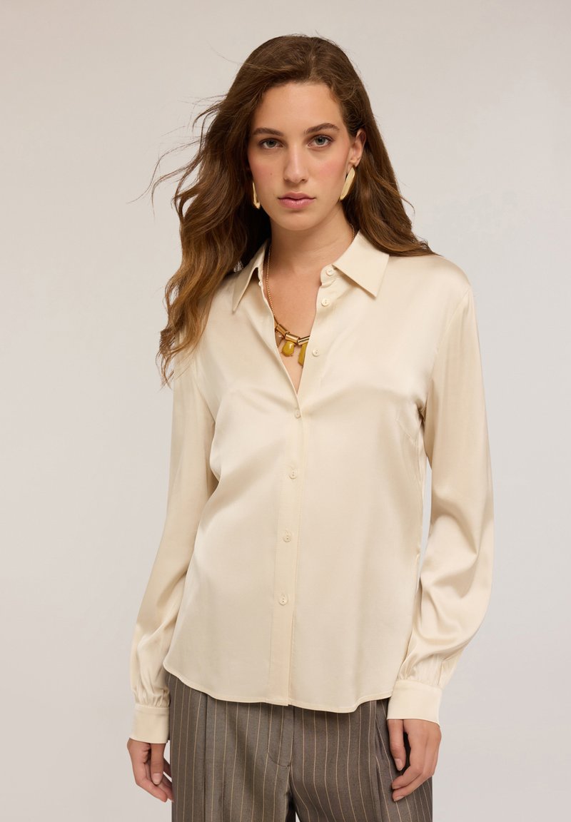 Donna con lunghi capelli mossi, che indossa una camicia di seta beige con bottoni, collana d'oro, orecchini d'oro e pantaloni a righe sottili, sullo sfondo chiaro.