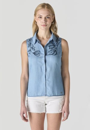 Mujer con camisa sin mangas azul claro abotonada con bordado floral negro y pantalones cortos blancos deshilachados, posando sobre un fondo liso.