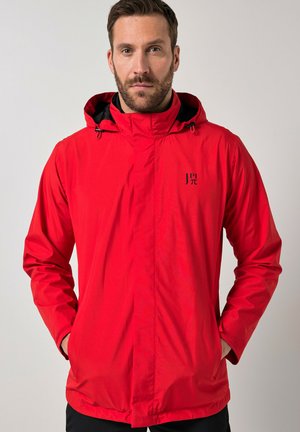 Mand med kort brunt hår og skæg iført en rød hættetrøje med hænderne i lommerne, stående mod en ensfarvet lys baggrund.