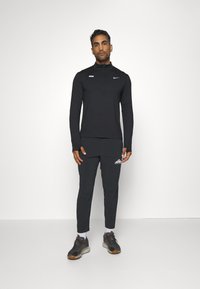 Schwarzes Sportoberteil mit hohem Kragen und Viertelreißverschluss, kombiniert mit schwarzen, schmal zulaufenden Hosen mit einem weißen Logo, abgerundet durch dunkle Sneakers.