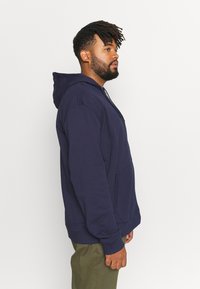 Sweat à capuche oversize bleu marine en mélange de coton, doté d'une poche avant, d'une capuche avec cordon et de poignets côtelés, porté avec un pantalon vert olive.