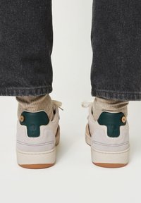 Baskets avec un corps en daim beige, accent en cuir vert sur le talon, semelle en caoutchouc marron et chaussettes tricotées beiges dépassant du col.