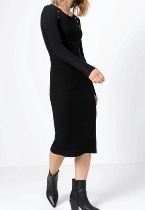 Robe noire ajustée à manches longues, tricot serré, col rond, œillets décoratifs aux épaules, associée à des bottines noires.