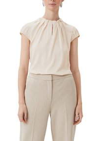 Short-sleeved, cream blouse with a gathered neckline, featuring a keyhole detail. Paired with light-colored, tailored trousers.