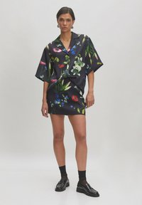 Zwarte bloemenjurk met grote, kleurrijke bloesems op een satijnachtige stof. Relaxte pasvorm en korte mouwen met een rechte zoom. Gemaakt met zwarte schoenen.