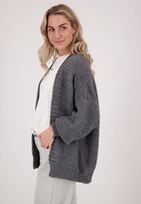 Grijze gebreide cardigan met een losse pasvorm, geribde manchetten en open voorkant, gedragen over een wit shirt en gestreepte broek. Bevat zijzakken.