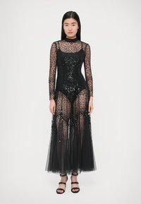 Black sheer maxi dress with long sleeves, embellished with sequins. Underlay bodysuit features a fitted design. Worn with black strappy heels.