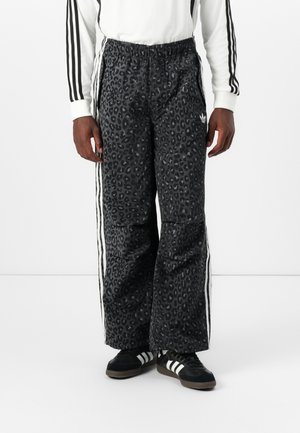 Person trägt schwarze, weit geschnittene Hose mit Leopardenmuster und weißen Seitenstreifen, schwarze Sneakers mit weißen Streifen und ein weißes Langarmshirt mit schwarzen Streifen.