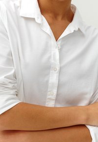 Chemise blanche à boutons avec col, présentant de longues manches retroussées, une texture de tissu lisse et de petits boutons blancs.