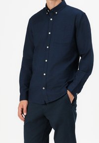 Camicia blu navy a maniche lunghe con bottoni, collo, tasca frontale e tessuto strutturato. Presenta bottoni bianchi e dettagli sui polsini.