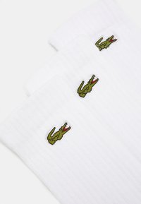Weiße gerippte Socken mit einem grünen gestickten Krokodil-Logo, das sich nahe der oberen Kante jeder Socke befindet. Weiche Textur, drei Paar sind abgebildet.
