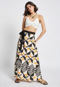 Maxi skirt featuring a geometric pattern in black, white, and orange with a tied black waistband. Model wears a white top and accessories.