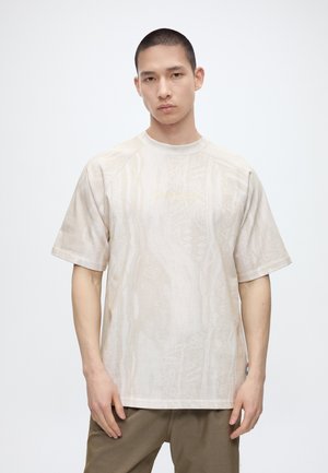 Mand med kort hår iført en løs, lysebeige mønstret T-shirt og olivengrønne bukser, stående mod en ensfarvet hvid baggrund.