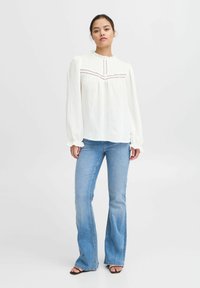 Witte blouse met lange mouwen en ruches bij de halslijn, met kantdetails, gecombineerd met lichtblauwe flared jeans en zwarte sandalen.