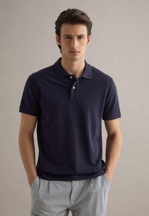 Giovane uomo con capelli scuri che indossa una polo blu scuro e pantaloni a righe bianche e blu, in piedi con le mani nelle tasche.