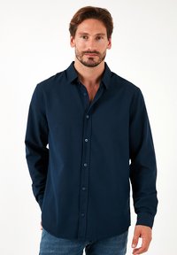 Camisa de botones azul marino en un tejido suave, con cuello estándar, mangas largas y dobladillo recto con botones azules visibles.