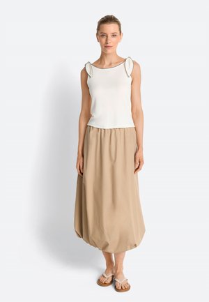 Frau trägt ein weißes ärmelloses Top mit Schulterdetails, einen beigen Ballonrock und beige Sandalen, steht vor einem schlichten Hintergrund.
