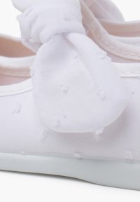 Weißer Stoffschuh mit strukturierten Punkten, ausgestattet mit einer Stoffschleife am Absatz und einer weißen Gummisohle.