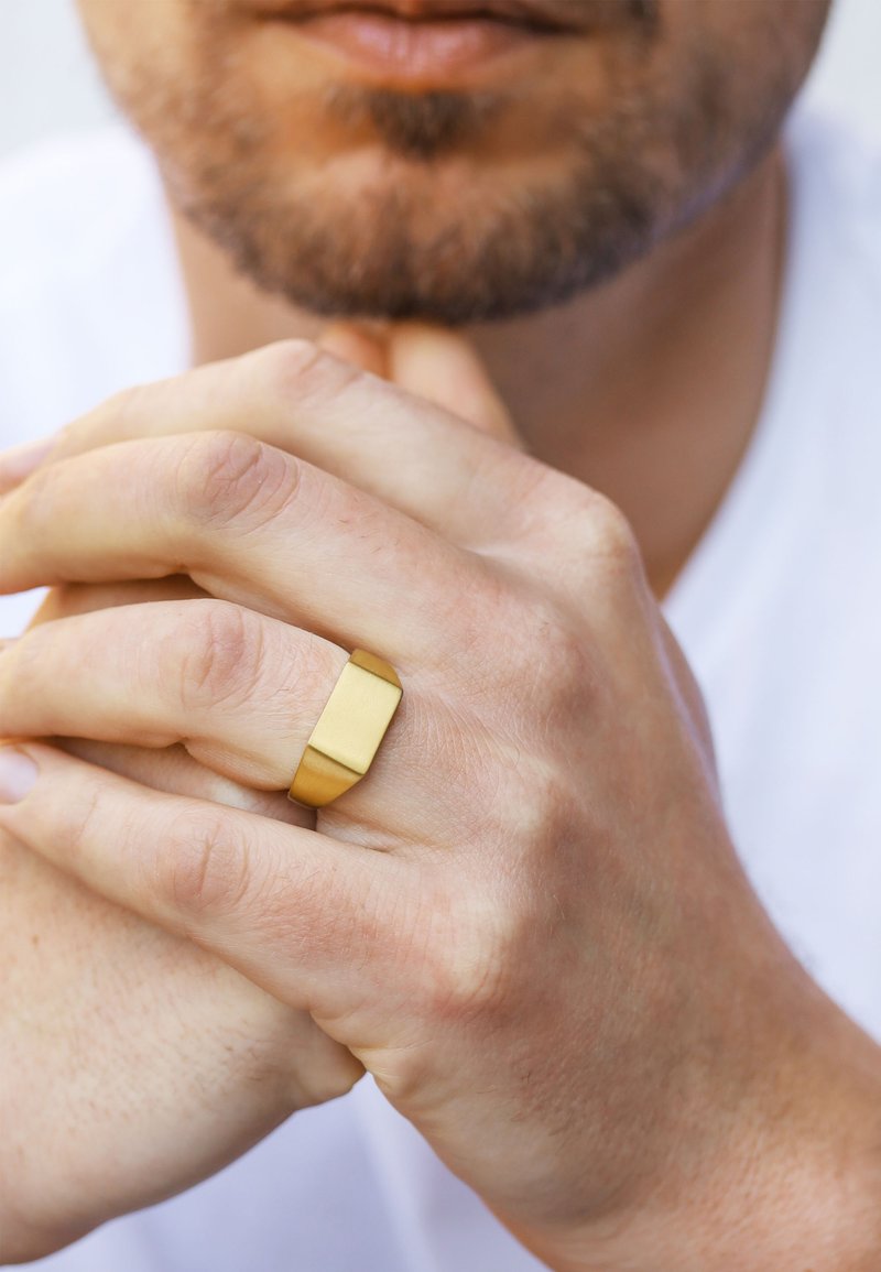 Matte goldfarbener Bandring mit rechteckigem Design, zwischen den Fingern gehalten. Haut sichtbar darunter, Hände entspannt und leicht strukturiert.