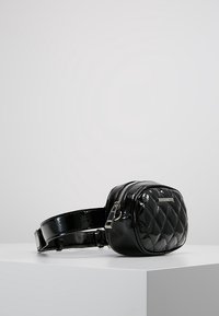 Piccola borsa a tracolla in pelle nera trapuntata con zip argentata e tracolla regolabile su una superficie bianca.