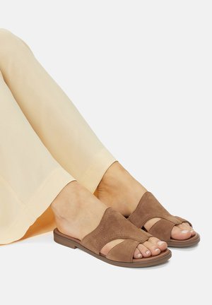 Sandale din suede maro, cu vârf deschis și design cu curea împletită, asortate cu pantaloni galbeni deschiși. Talpă plată, stil minimalist.