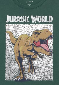 Zöld, rövid ujjú póló, amelynek elülső oldalán egy nagy barna T-Rex grafika található, fehér és ezüst textúrájú háttérrel, valamint a "Jurassic World" felirat.